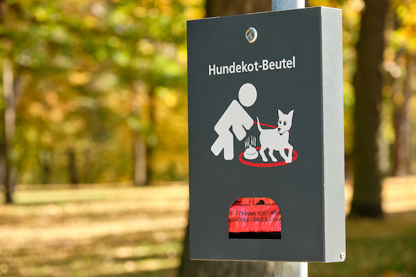 Grey dispenser with red “ Hundekot-Beutel “ (what means dog waste bag / dog excrement bag in English) in front of a park with autumn trees in Germany
